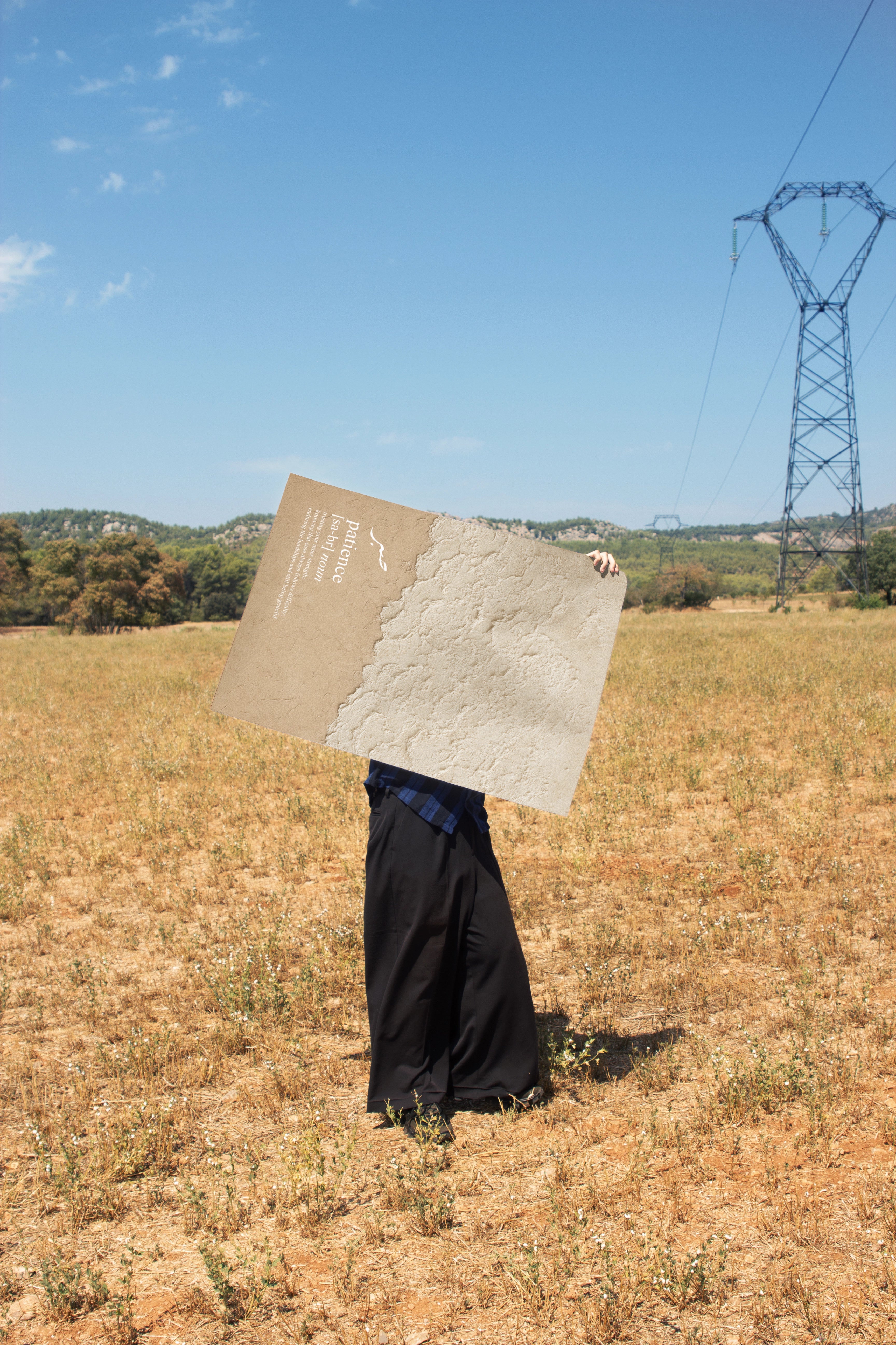 Sükür Poster mit beiger Strukturfläche und kalligrafischem Text, Person hält grossen Kunstdruck in trockener Landschaft vor blauem Himmel