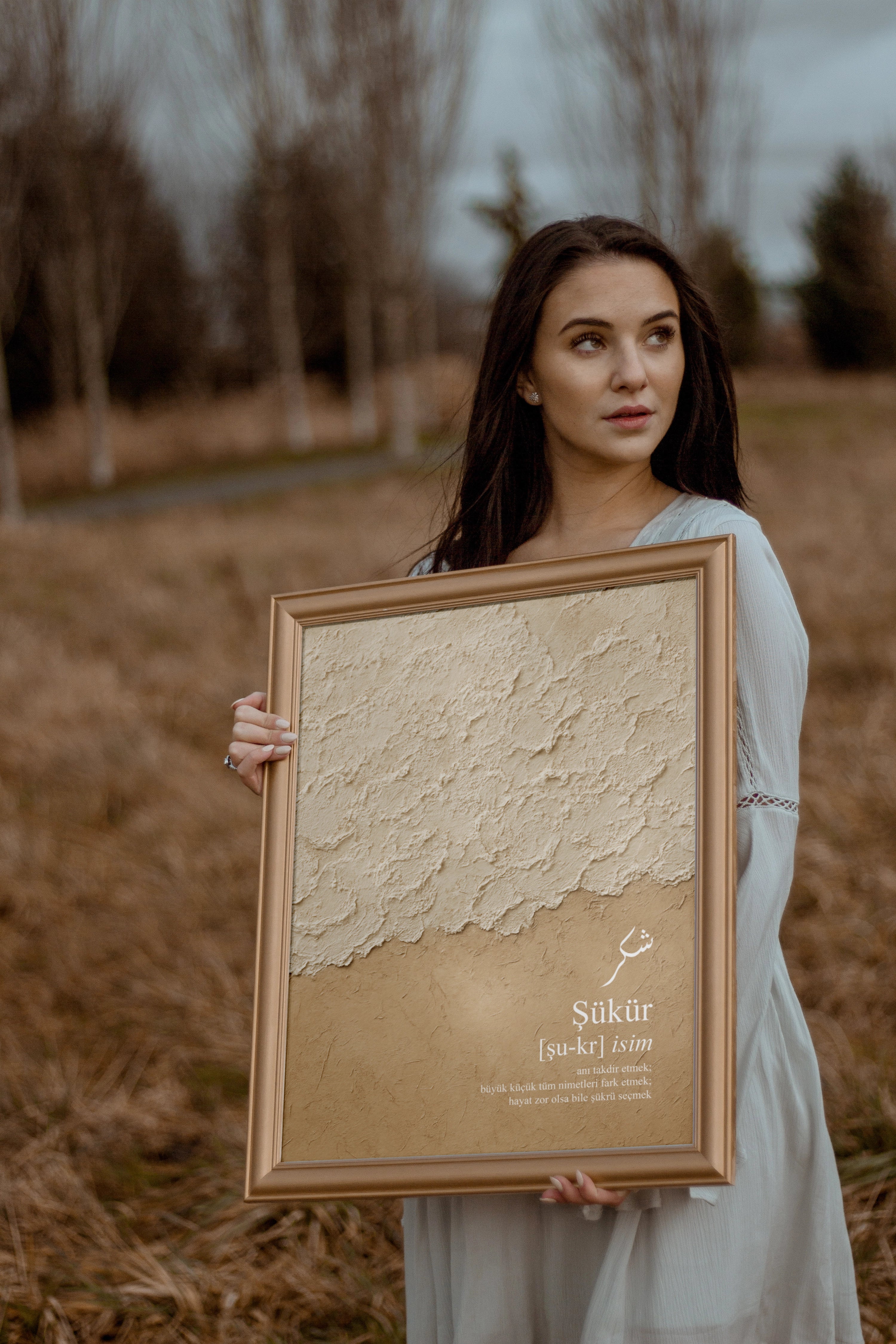 Sukur Poster in Goldrahmen mit kalligrafischem Schriftzug und Definition, junge Frau hält Wandbild in naturfarbener Landschaft
